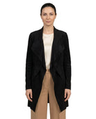 A woman modeling a black open-front wrap-style coat over a cream top and beige trousers, photographed against a plain white studio background.