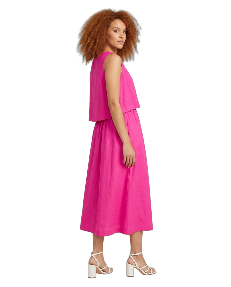 A woman with curly red hair is shown from the side wearing a bright pink sleeveless midi dress with a layered top and white strappy high-heeled sandals, set against a plain white background.