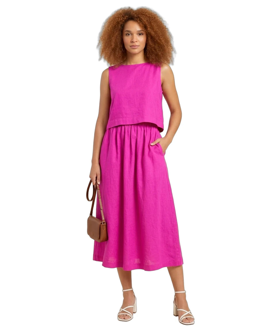 A woman modeling a bright pink two-piece linen set consisting of a sleeveless cropped top and a matching midi skirt with pockets, styled with a brown handbag and white strappy heels against a white background.