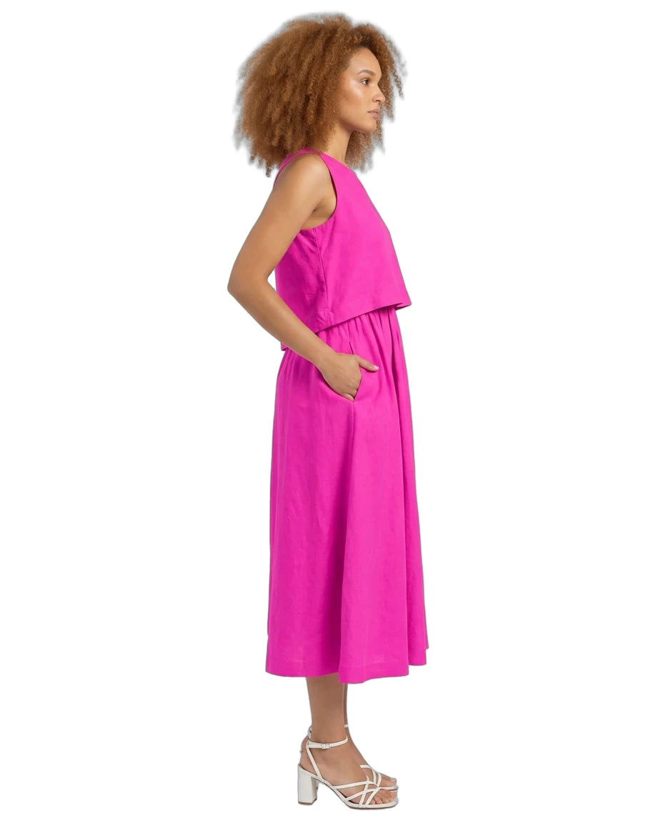 A woman shown in side profile wearing a sleeveless bright pink midi dress with a draped yoke and side pockets, paired with white strappy high-heeled sandals, against a plain white background.