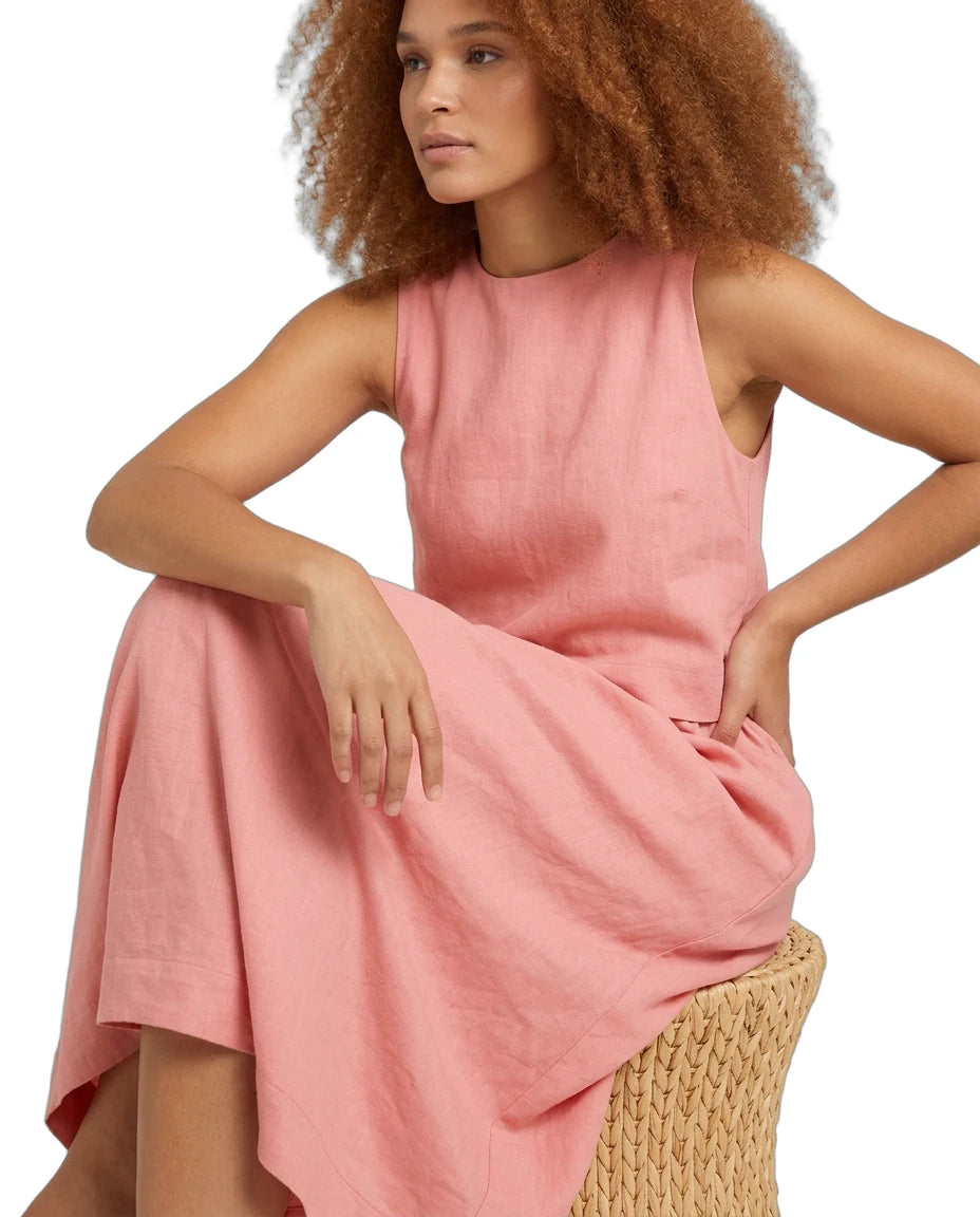 A woman with curly auburn hair wearing a sleeveless dusty pink linen dress, sitting on a woven straw chair against a white background.