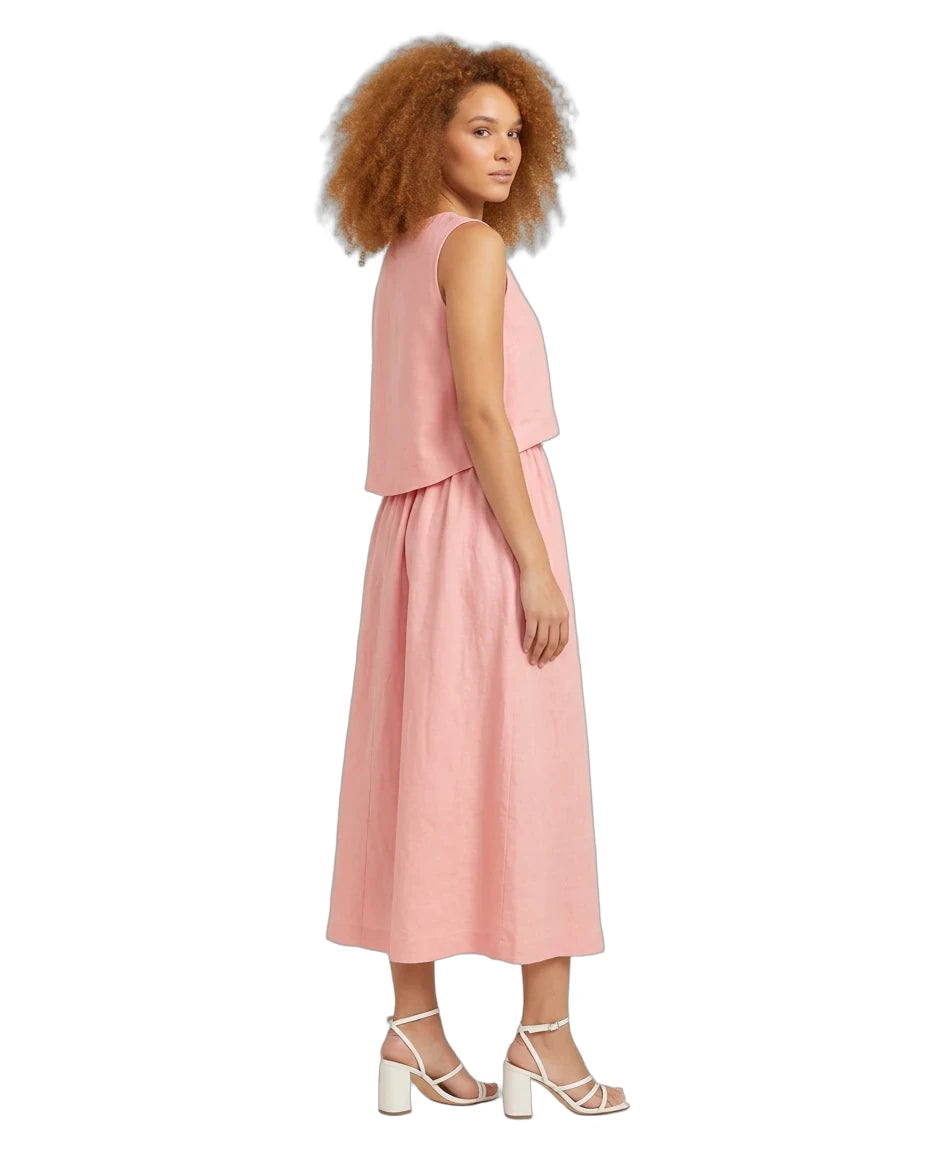 A woman with curly light brown hair is shown from the side wearing a sleeveless dusty pink midi dress with a layered ruffle top and white strappy high-heeled sandals, set against a plain white background.