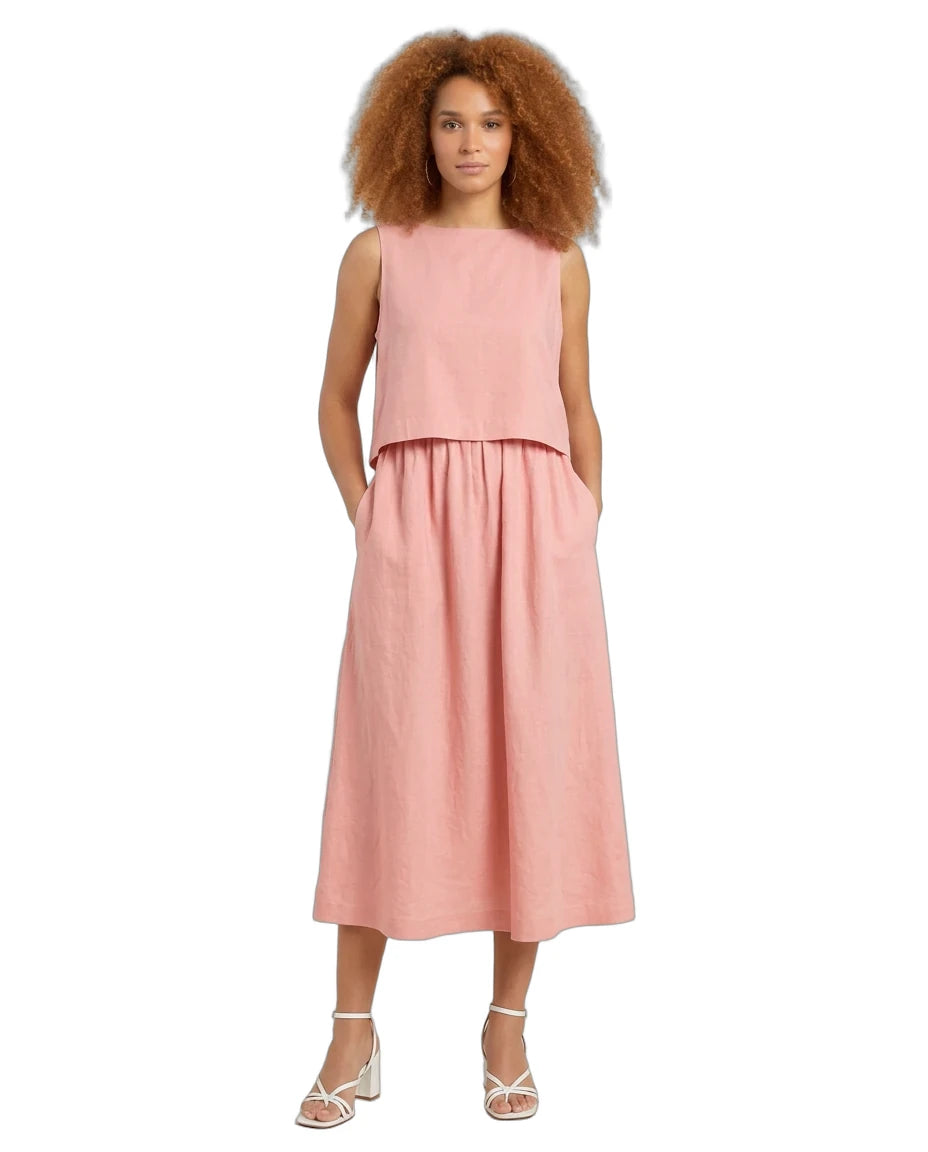 A woman with voluminous curly brown hair wearing a sleeveless dusty pink two-piece linen set consisting of a cropped top and a matching midi skirt with pockets, paired with white strappy high-heeled sandals, set against a plain white background.