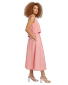 A woman with curly light brown hair is shown in profile wearing a sleeveless dusty pink linen midi dress with a ruffled overlay and side pockets, paired with white strappy block-heel sandals, against a solid white background.