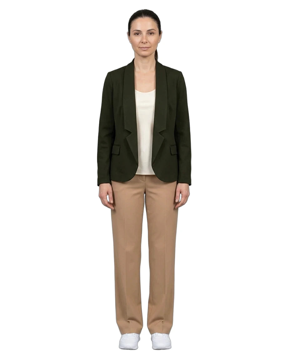 A woman stands against a plain white background wearing a dark olive green blazer over a cream top, beige trousers, and white sneakers.