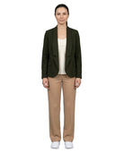 A woman stands against a plain white background wearing a dark olive green blazer over a cream top, beige trousers, and white sneakers.