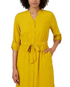 A woman wearing a mustard yellow button-front shirt dress with a tie waist and rolled sleeves, shown against a plain white background.