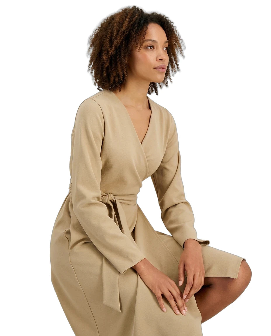 A woman with curly brown hair wearing a beige wrap dress with long sleeves and a tie waist, posing against a solid white background.