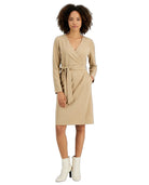 A woman modeling a beige wrap dress with long sleeves and a matching waist tie, paired with white ankle boots, against a plain white background.