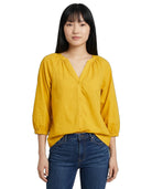 A woman wearing a mustard yellow blouse with three-quarter puff sleeves and a gathered V-neckline, paired with dark blue jeans, against a plain white background.