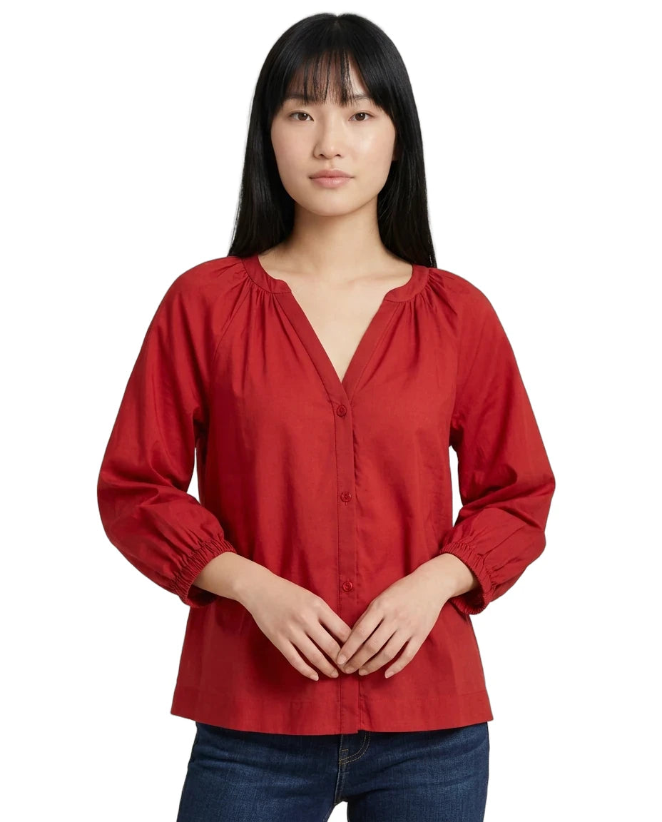 A woman wearing a red long-sleeve blouse with a split V-neckline, gathered shoulders, and button-front closure, paired with dark blue jeans against a plain white background.