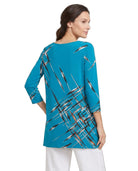 A woman shown from the back wearing a turquoise blue tunic top with a black, white, and gray brushstroke pattern, paired with white pants, against a plain white background.