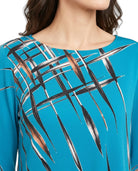 A close-up of a model wearing a bright turquoise blue top with a black, white, and brown abstract leaf-like pattern across the chest.