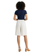 A woman shown from the back wearing a navy blue short-sleeve top, off-white pleated knee-length shorts, and tan block-heel mules against a plain white background.
