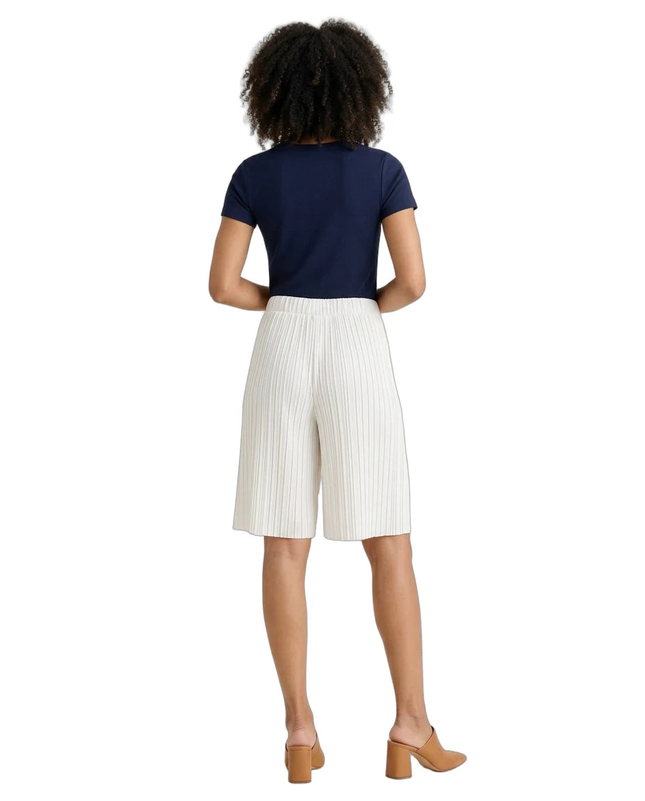 A woman shown from the back wearing a navy blue short-sleeve top, off-white pleated knee-length shorts, and tan block-heel mules against a plain white background.