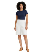 A woman modeling a navy blue short-sleeve crewneck top and white pleated knee-length shorts, paired with tan heeled mules, against a plain white background.