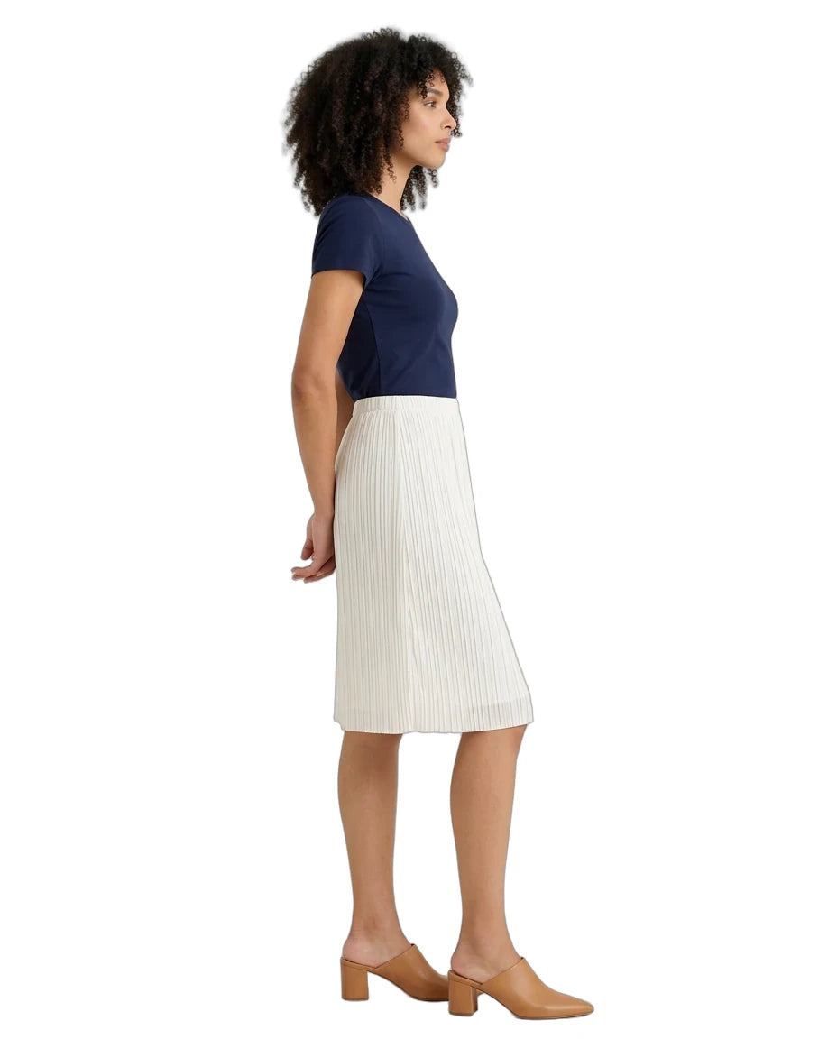 A woman shown in profile wearing a navy blue short-sleeve top and a white pleated midi skirt, paired with tan block-heel mules, against a plain white background.