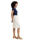 A woman shown in profile wearing a navy blue short-sleeve top and a white pleated midi skirt, paired with tan block-heel mules, against a plain white background.
