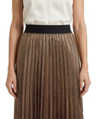 A close-up of a model wearing a pleated metallic bronze skirt with a wide black waistband, paired with a cream top against a white background.