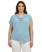 Woman wearing a light blue cowl-neck top and white pants on a white background