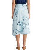 A model is shown from the back wearing a light blue midi skirt with a dark blue abstract paint splatter pattern, paired with a navy top and tan block-heel sandals, against a plain white background.