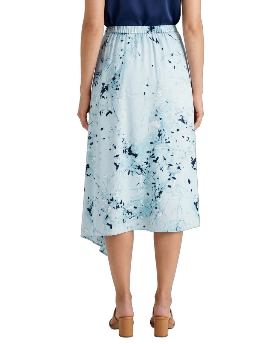 A model is shown from the back wearing a light blue midi skirt with a dark blue abstract paint splatter pattern, paired with a navy top and tan block-heel sandals, against a plain white background.