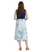A woman viewed from the back wearing a sleeveless dress with a navy blue bodice and a light blue watercolor-style printed skirt, paired with tan block-heel sandals on a white background.