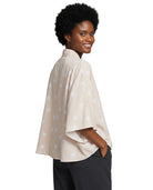 A woman with short black hair is shown from the back-three-quarter view wearing a light beige cape-style top with a subtle white star pattern, paired with dark gray pants, against a plain white background.