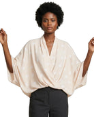 A woman modeling a light beige wrap-style blouse with a white dotted pattern and wide draped sleeves, paired with dark charcoal pants, against a plain white background.