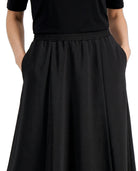 A close-up of a person wearing a black midi skirt with an elastic waistband and side pockets, paired with a black short-sleeve top, against a plain white background.