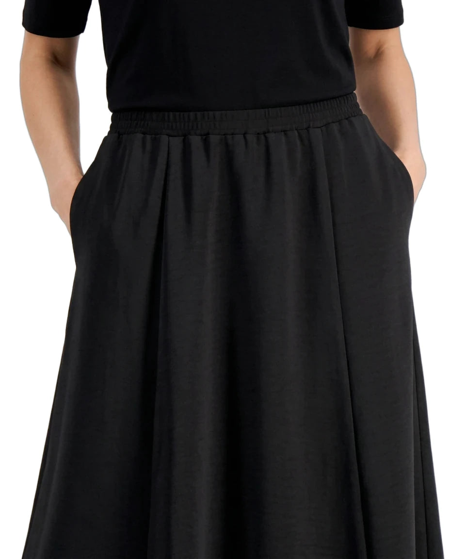 A close-up of a person wearing a black midi skirt with an elastic waistband and side pockets, paired with a black short-sleeve top, against a plain white background.