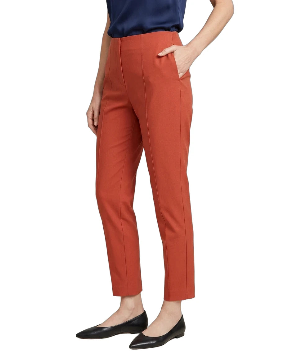 A model wearing a pair of cropped, straight-leg trousers in a warm rust orange color, paired with a navy top and black flats, shown from the waist down on a white background.