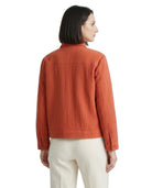 A woman shown from the back wearing a textured rust-orange quilted jacket and cream-colored trousers, photographed against a solid white background.