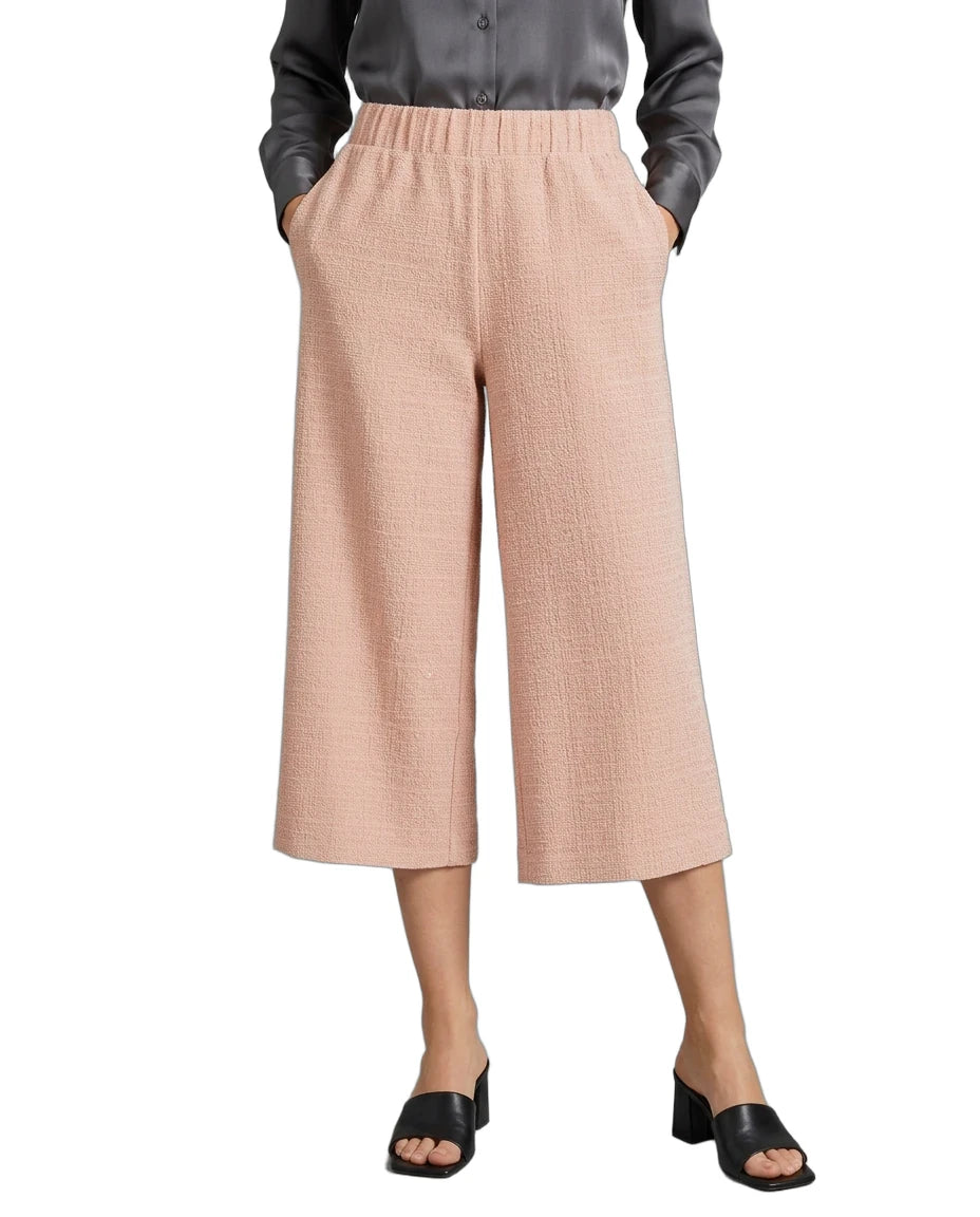 A model wearing a pair of light peach-pink cropped wide-leg pants with a textured weave, paired with a dark gray button-up shirt and black slide sandals, shown against a plain white background.
