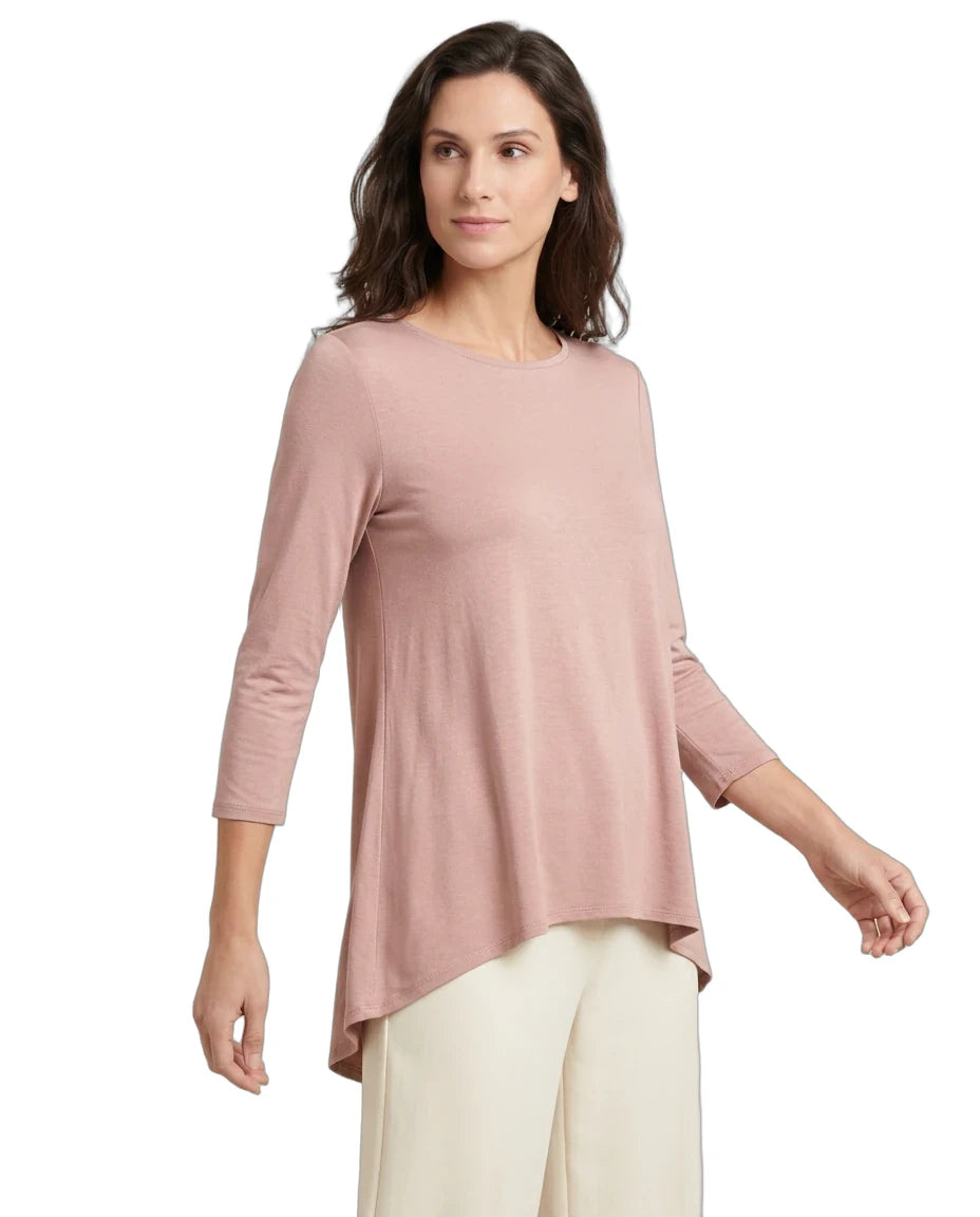A woman modeling a dusty pink three-quarter sleeve top with an asymmetrical handkerchief hem, paired with cream pants, against a plain white background.