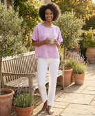 Woman in a pink shirt and white pants standing in a garden holding a mug.