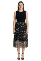 Woman wearing a black sleeveless top and a sheer black skirt with gold polka dots on a white background