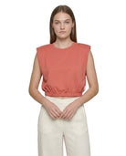 Woman wearing a coral sleeveless top and white pants on a white background
