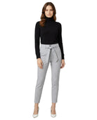 Woman wearing a black turtleneck and gray Calin Klein pants with a belt on a white background