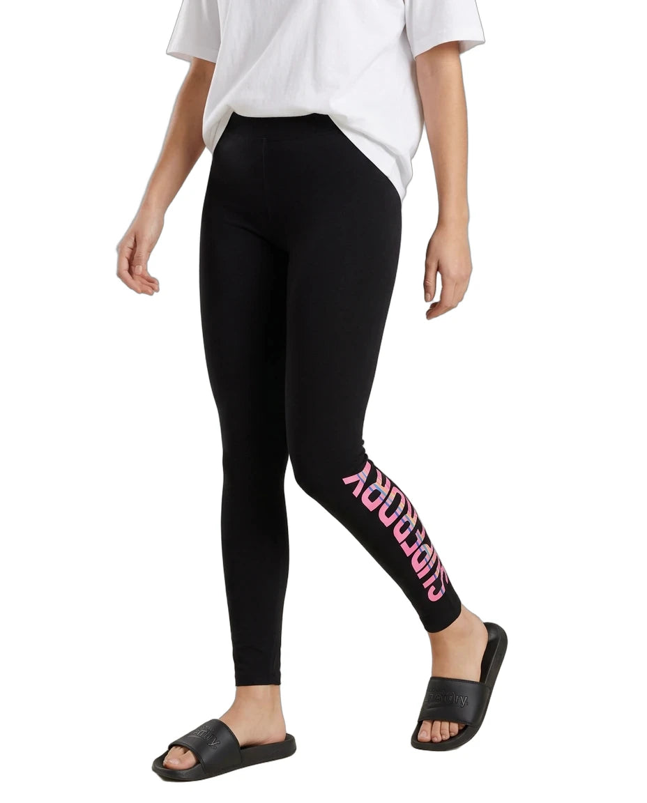 A model wearing black leggings with a pink 'SUPERDRY' logo printed vertically down the left leg, styled with a white t-shirt and black slide sandals against a plain white background.