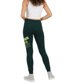 A model from the back wearing dark green heathered leggings with a bright yellow-green graphic text print on the left leg, paired with a white crop top and white sneakers, against a plain white background.