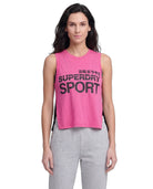 A woman modeling a bright pink sleeveless crop top with black 'SUPERDRY SPORT' text on the front, paired with light gray sweatpants, against a plain white background.