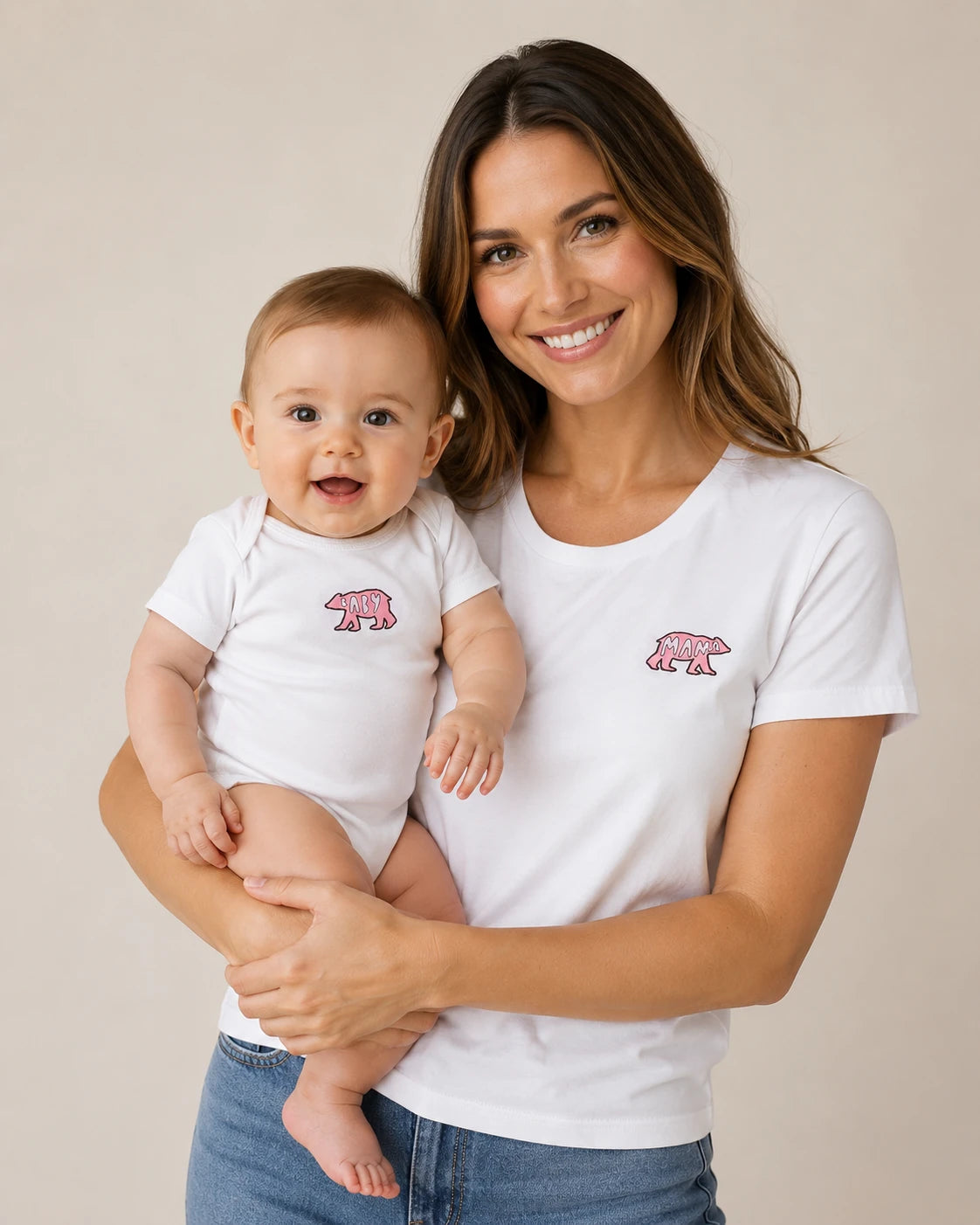 A smiling woman holding a baby, both wearing matching white short-sleeve bodysuits with a small pink polar bear graphic on the chest. The woman is also wearing blue jeans, and the background is a clean, neutral beige studio backdrop.