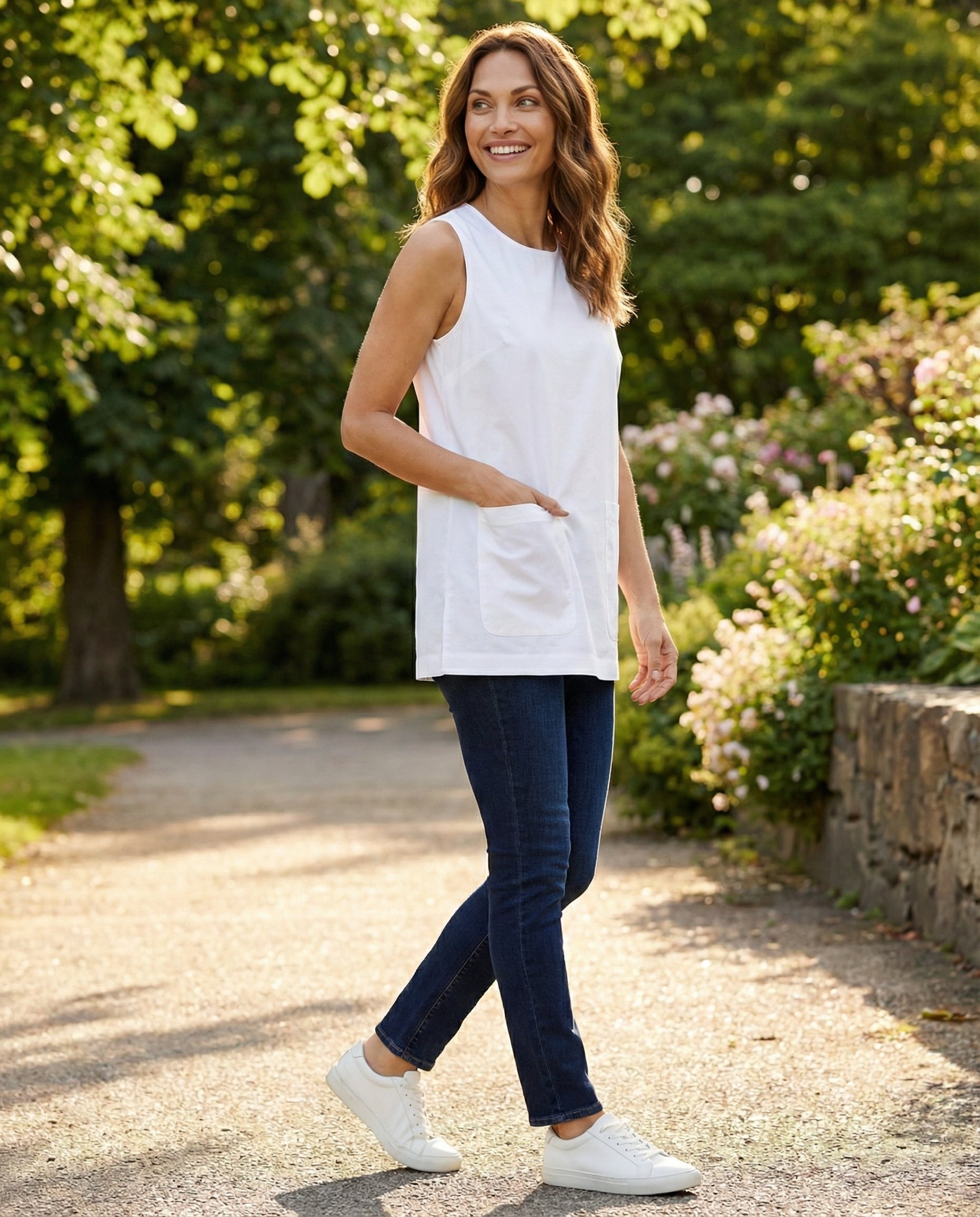 Woman in a white sleeveless top and blue jeans walking outdoors in a garden.